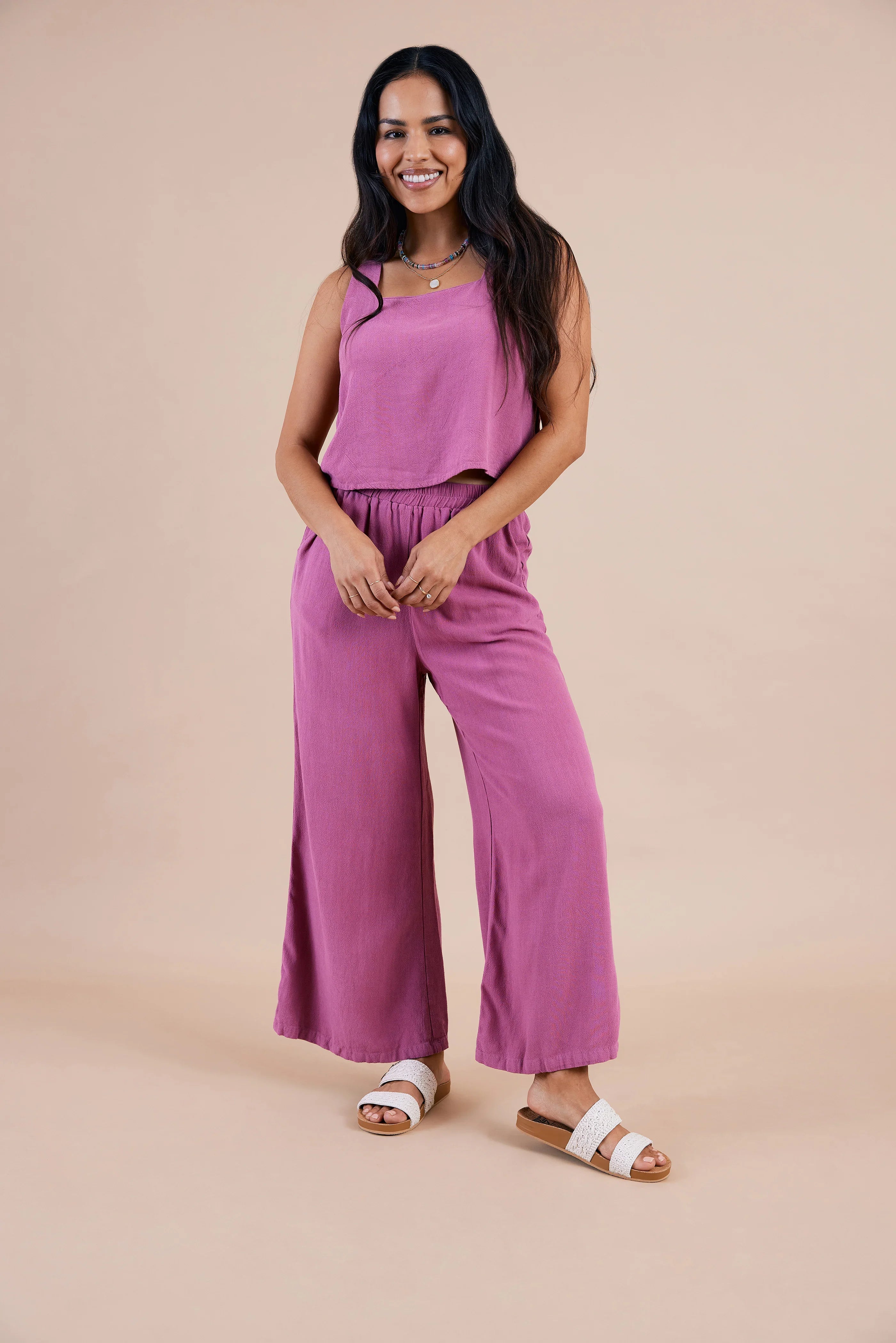 אישה לובשת סט טופ ומכנסיים רחבים בצבע ורוד, עומדת ומחייכת על רקע אחיד