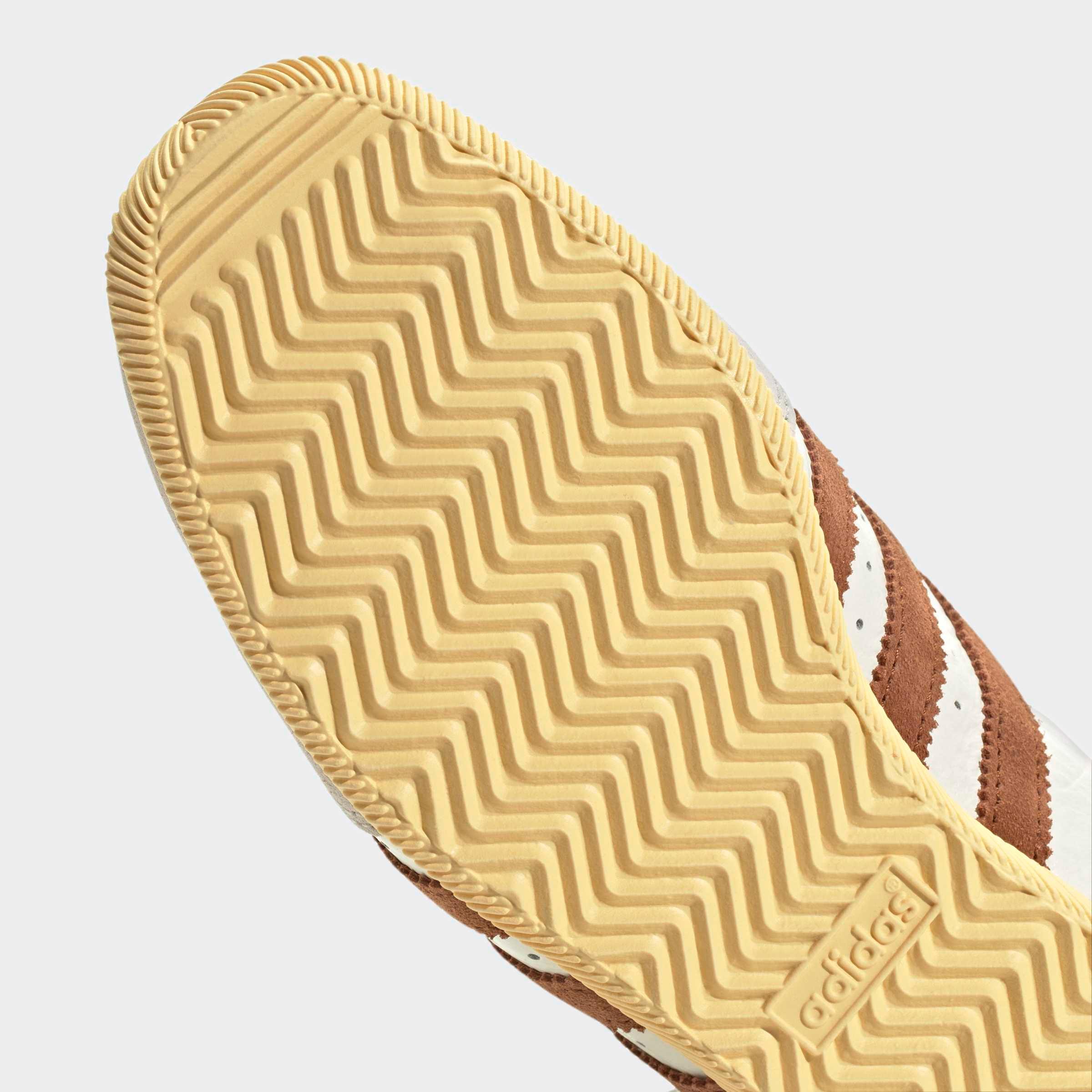 תקריב סוליית נעל ספורט עם דפוס זיגזג וצד חום עם פסים לבנים, רקע אפור