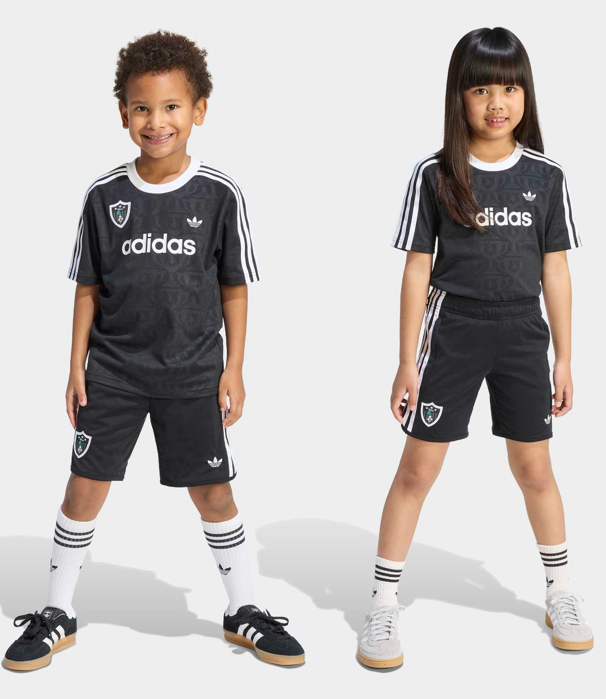 שני ילדים לובשים סט כדורגל שחור עם פסי adidas לבנים, חולצה קצרת שרוול ומכנס קצר