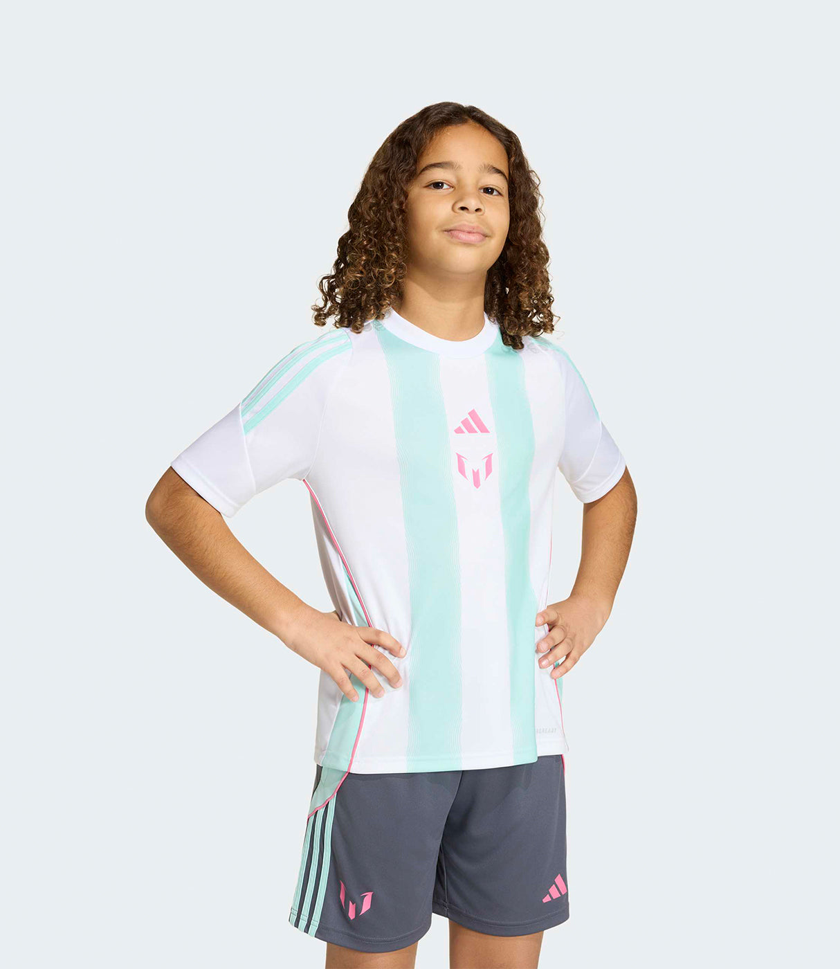 ילד לובש חולצת כדורגל לבנה עם פסים ירוקים ומכנסי ספורט אפורים, צילום על רקע חלק