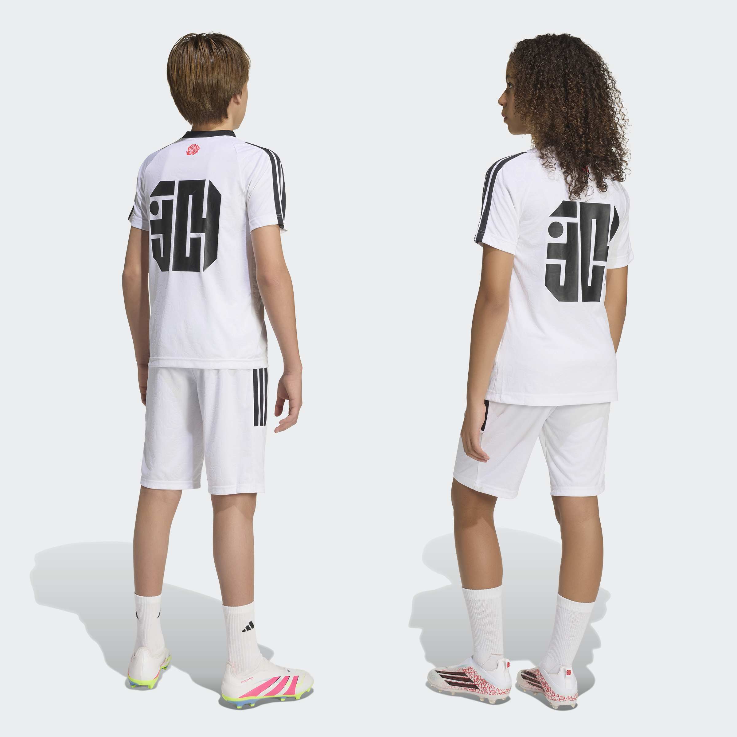 שני ילדים עם סט חולצת ושורט ספורט לבן, הדפס גרפי שחור בגב, נעלי כדורגל לבנות, מבט מאחור