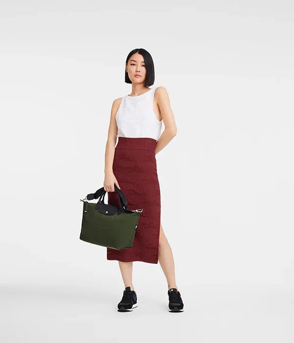 אישה לובשת חצאית בורדו, גופיה לבנה ונעלי ספורט, מחזיקה תיק ירוק מבית קבוצת אלפא CHOZEN