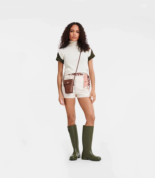 אישה עם שיער מתולתל לובשת חולצה לבנה, מכנסיים קצרים, מגפיים ירוקים ותיק צד חום, רקע לבן, מותגי אופנה CHOZEN