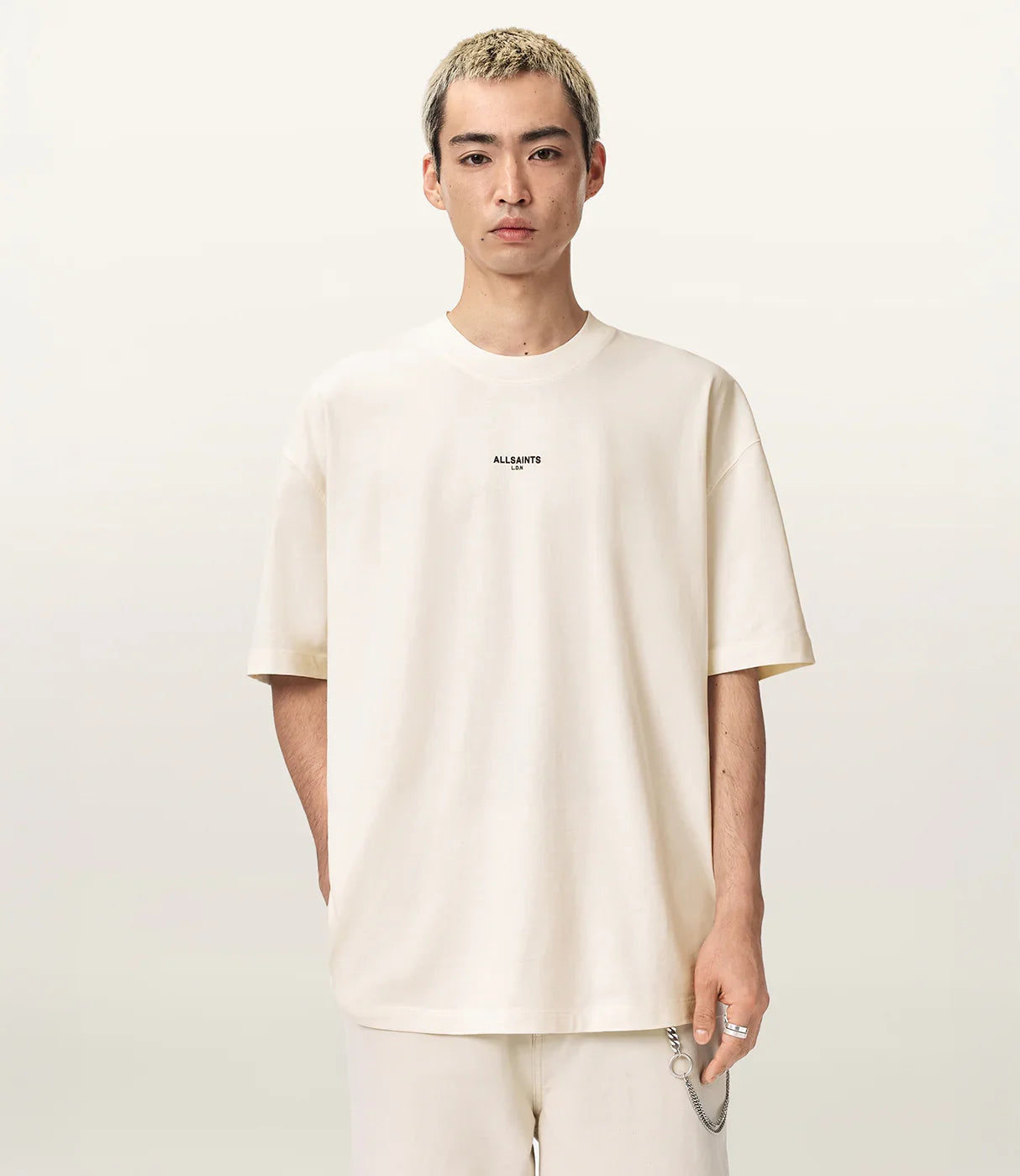 גבר לובש חולצת טי שרט בגוון שמנת עם הדפס ALLSAINTS במרכז החזה, מכנסיים שמנת, רקע ניטרלי