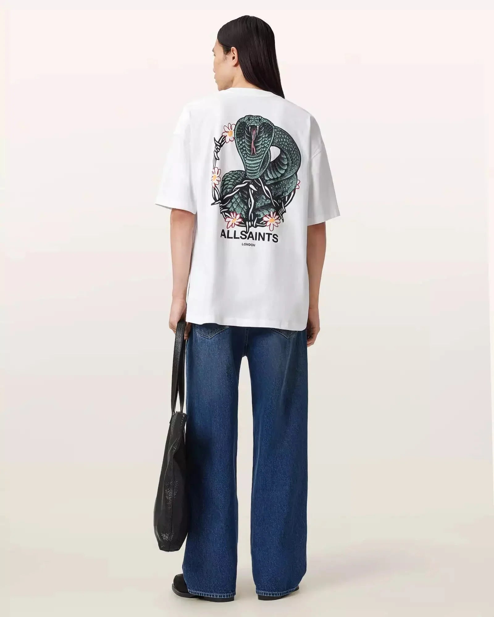 אישה עם שיער ארוך לובשת חולצת טי לבנה של ALLSAINTS עם הדפס נחש ופרחים, וג'ינס רחב מבית CHOZEN
