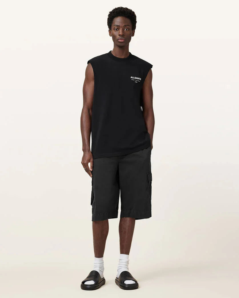 אופנת גברים CHOZEN: גבר לובש חולצת שרוולים שחורה ומכנסי ברמודה שחורים, נעלי סליפר וגרביים לבנים