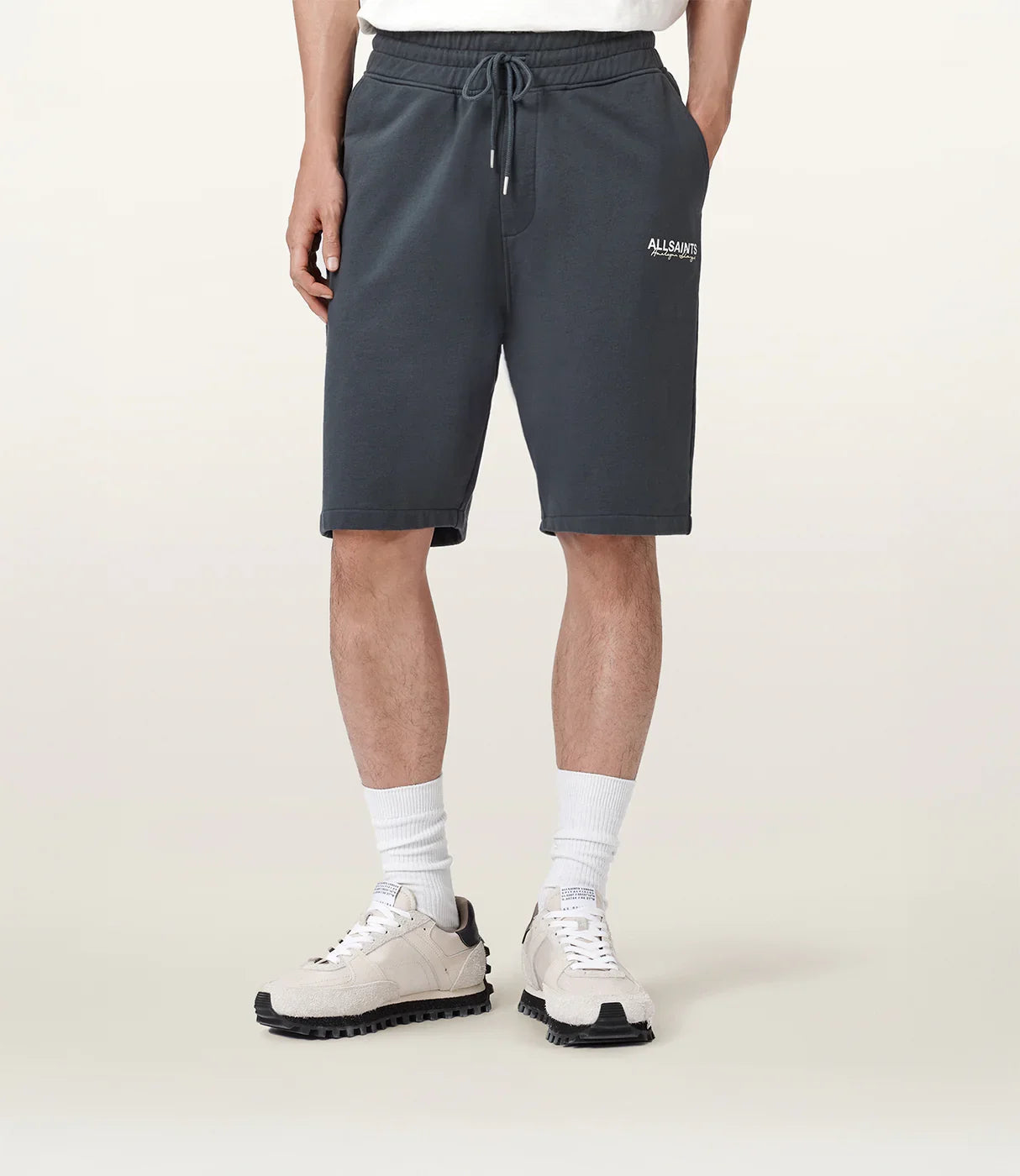 גבר לובש מכנסי ברמודה ספורט כהים עם מותג, חולצה לבנה, גרביים ונעליים בהירות