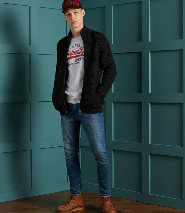 גבר לובש ג'ינס כחול, חולצת טי אפורה עם הדפס, סוודר שחור וכובע משובץ, מצולם לרקע כחול, אופנת CHOZEN