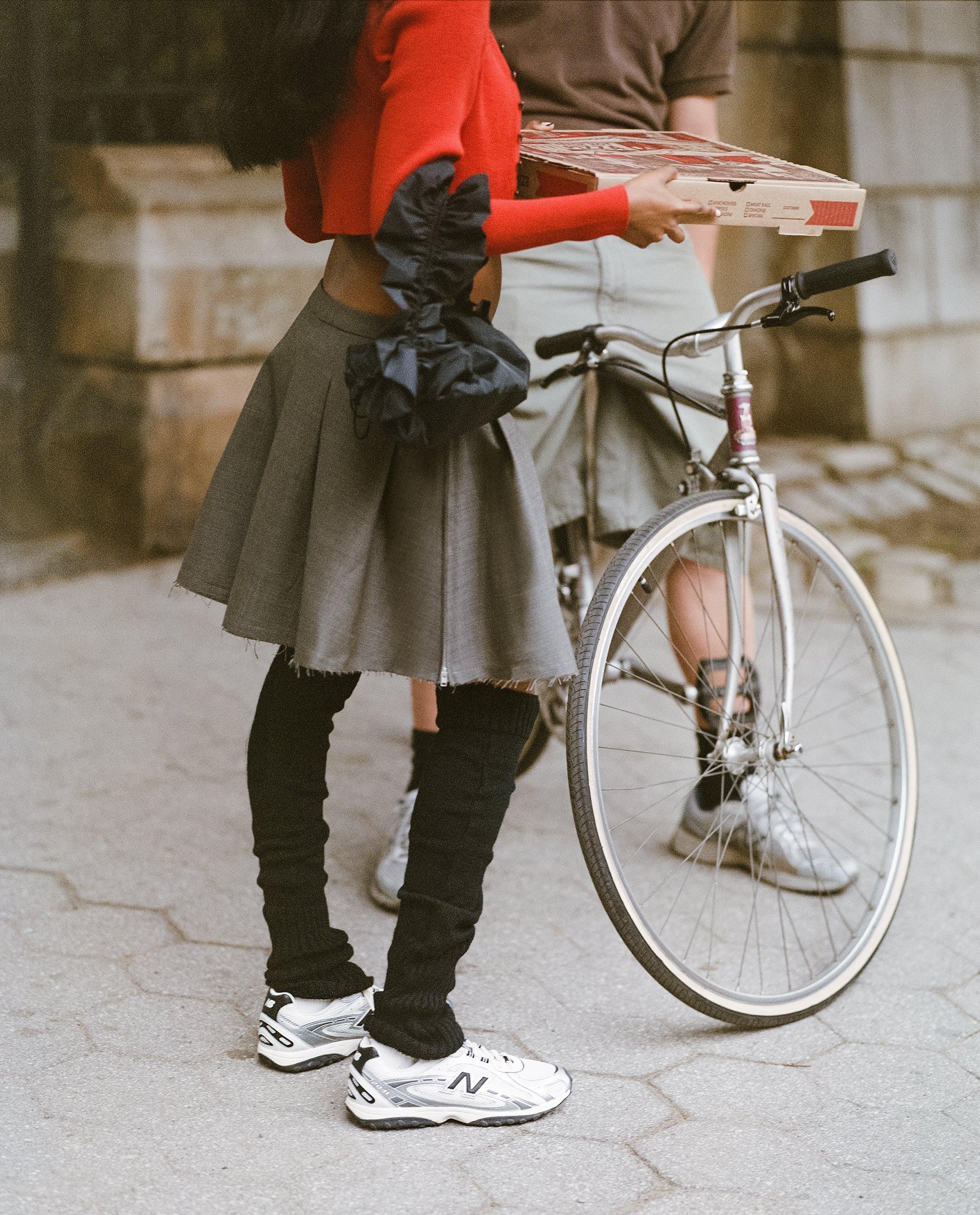 אישה עם סווטשירט אדום, חצאית אפורה ונעלי ניו באלאנס, ליד אופניים וקופסת פיצה, אופנת רחוב CHOZEN