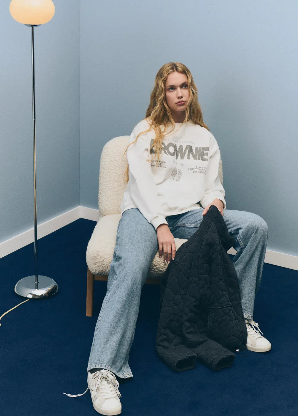 אופנת נשים של CHOZEN: דוגמנית עם סווטשרט BROWNIE, מכנסי ג'ינס ונעלי ספורט, מצולמת בחדר מודרני