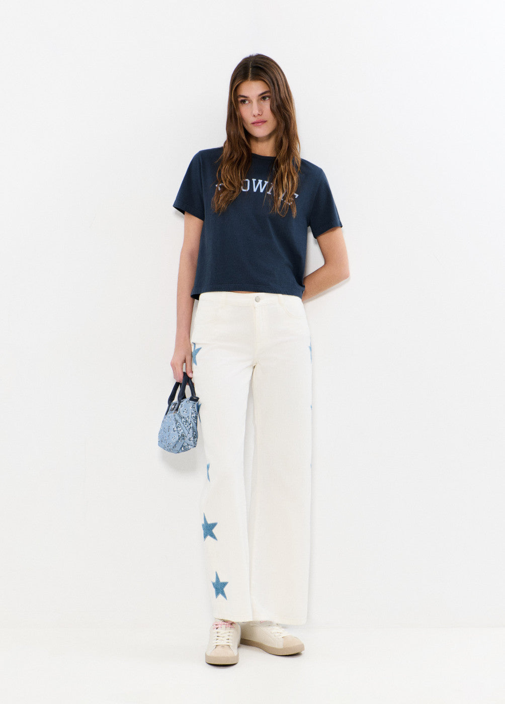 אישה עם טי שירט כחולה, מכנסי כותנה לבנים עם כוכבים כחולים, מחזיקה תיק קטן