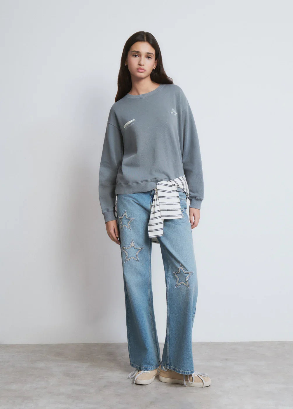 דוגמנית לובשת סווטשירט אפור, ג'ינס עם עיטורי כוכבים ונעליים שטוחות – אופנה טרנדית CHOZEN
