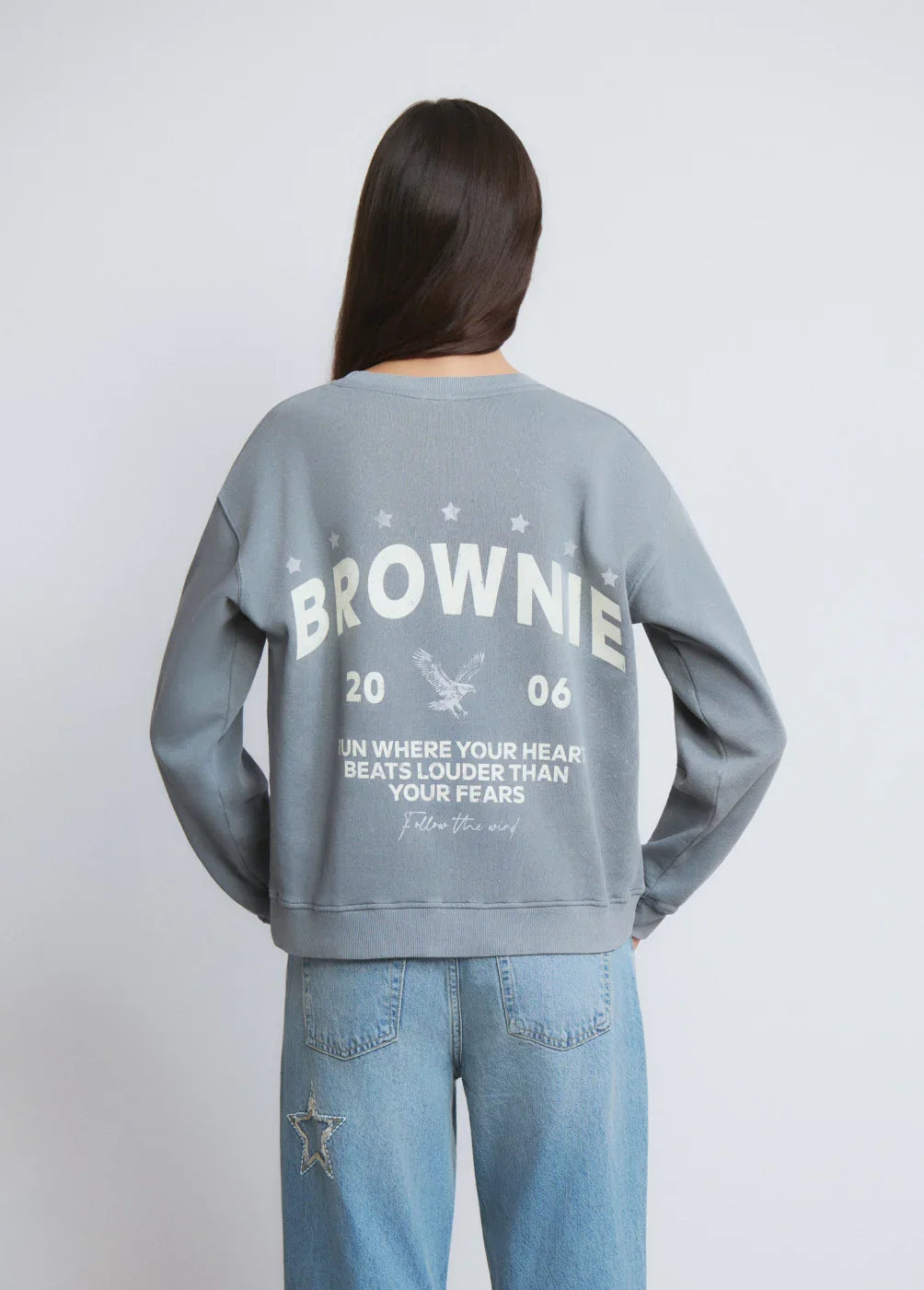 אישה בלבוש קז'ואל עם סווטשירט אפור של BROWNIE וכיתוב גב, מכנסי ג'ינס עם כוכב, CHOZEN