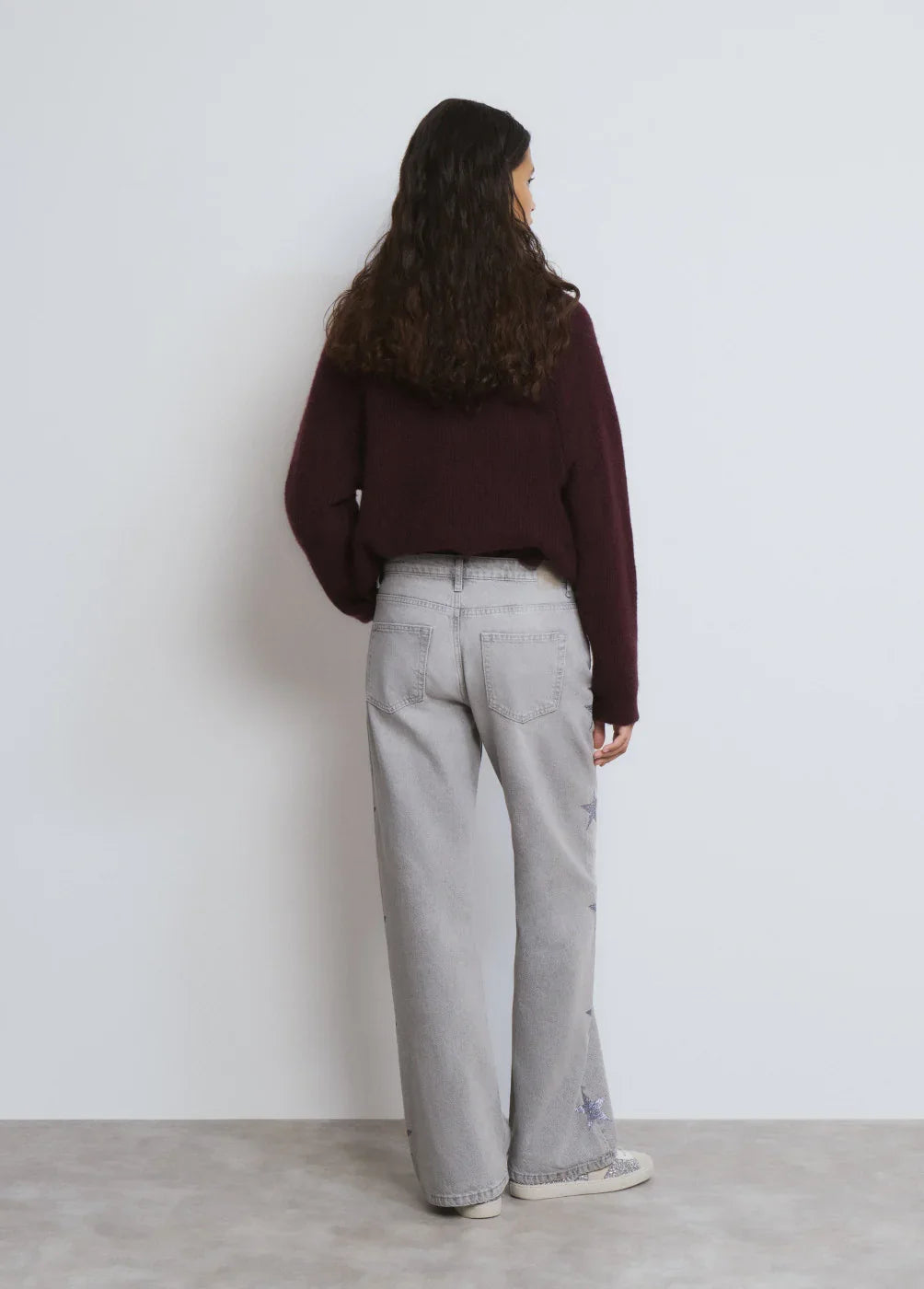 אישה עם סוודר בורדו ומכנסי ג'ינס אפורים עם כוכבים, עומדת בחזית לבנה, CHOZEN