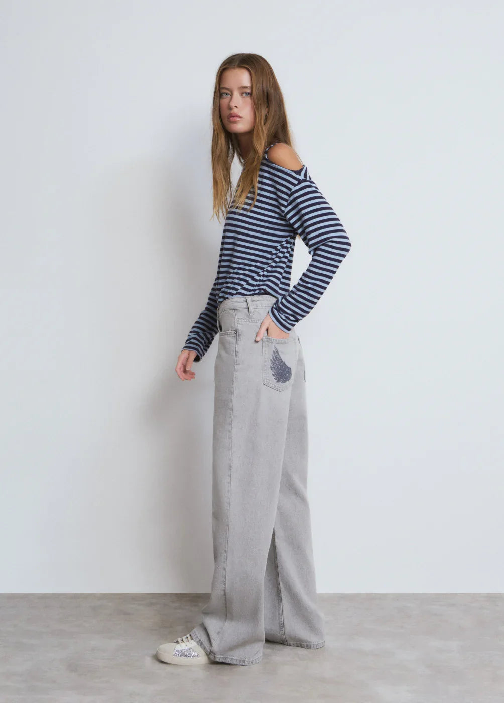דוגמנית עם חולצה פסים כחולה ומכנסי ג'ינס רחבים אפורים, אופנת CHOZEN, רקע לבן