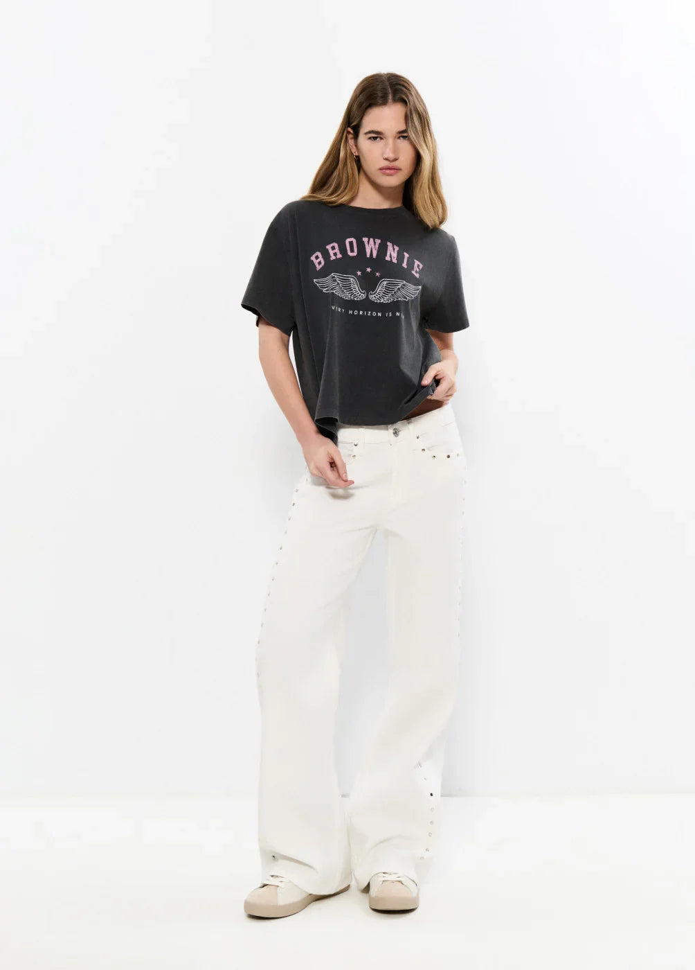 אישה לובשת חולצת טי שחורה עם כיתוב BROWNIE ומכנסיים לבנים רחבים, סטודיו מינימליסטי