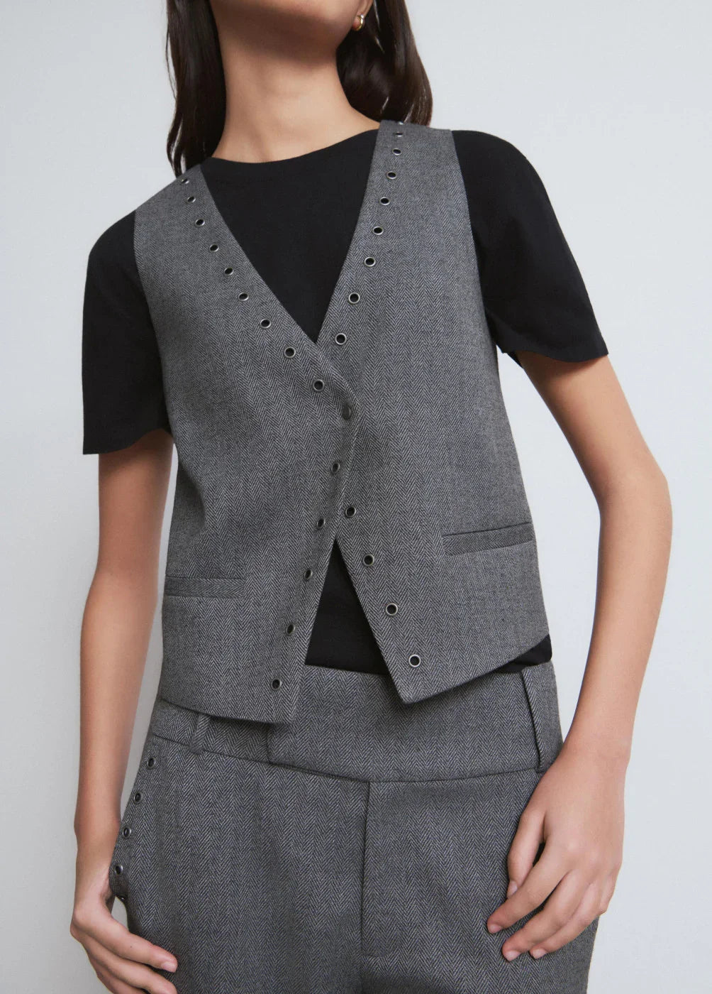 אישה לובשת וסט מחויט אפור עם ניטים ומכנסיים תואמים, חולצת טי שחורה מתחת