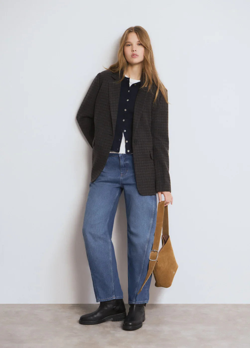 דוגמנית עומדת עם בלייזר כהה, סוודר כחול, ג'ינס ונעליים שחורות, מחזיקה תיק חום מבית CHOZEN