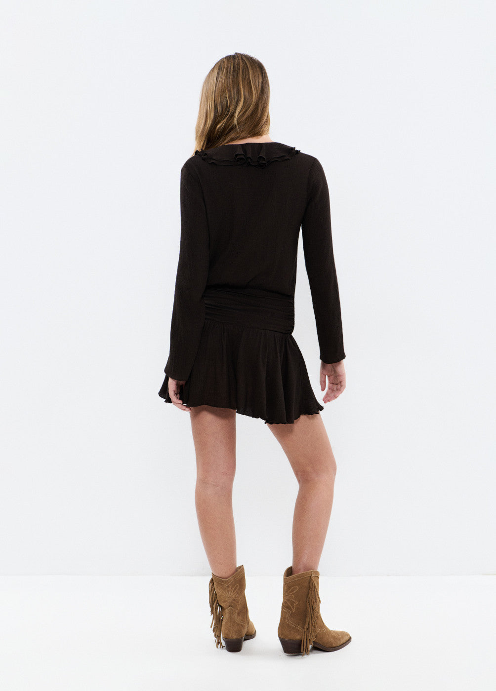 אישה לובשת שמלה שחורה עם שרוולים ארוכים ומגפיים חומים, מצולמת מאחור על רקע לבן