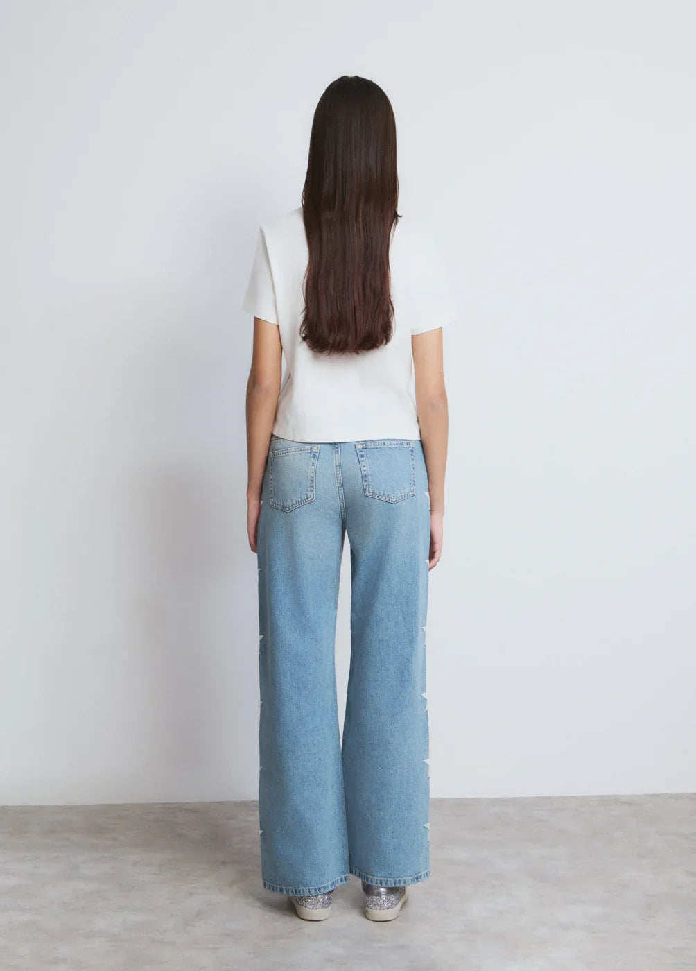 אישה עם שיער ארוך לובשת ג'ינס רחב וחולצה לבנה, צילום מאחור על רקע קיר לבן, CHOZEN