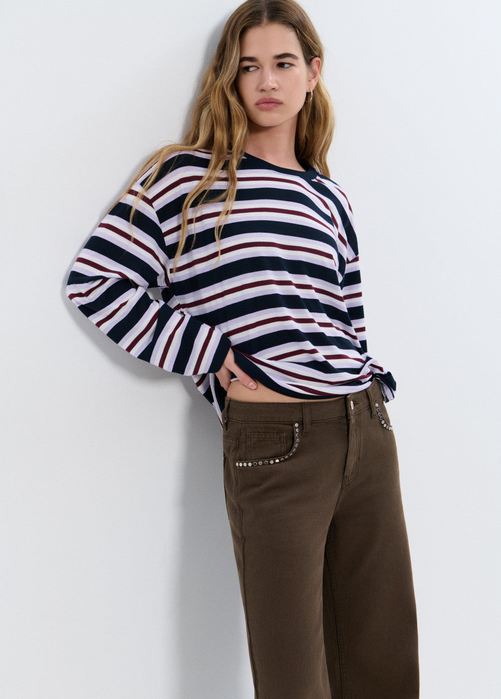 אישה לובשת חולצת פסים ארוכה בצבעי כחול, לבן ובורדו ומכנסיים חומים עם ניטים