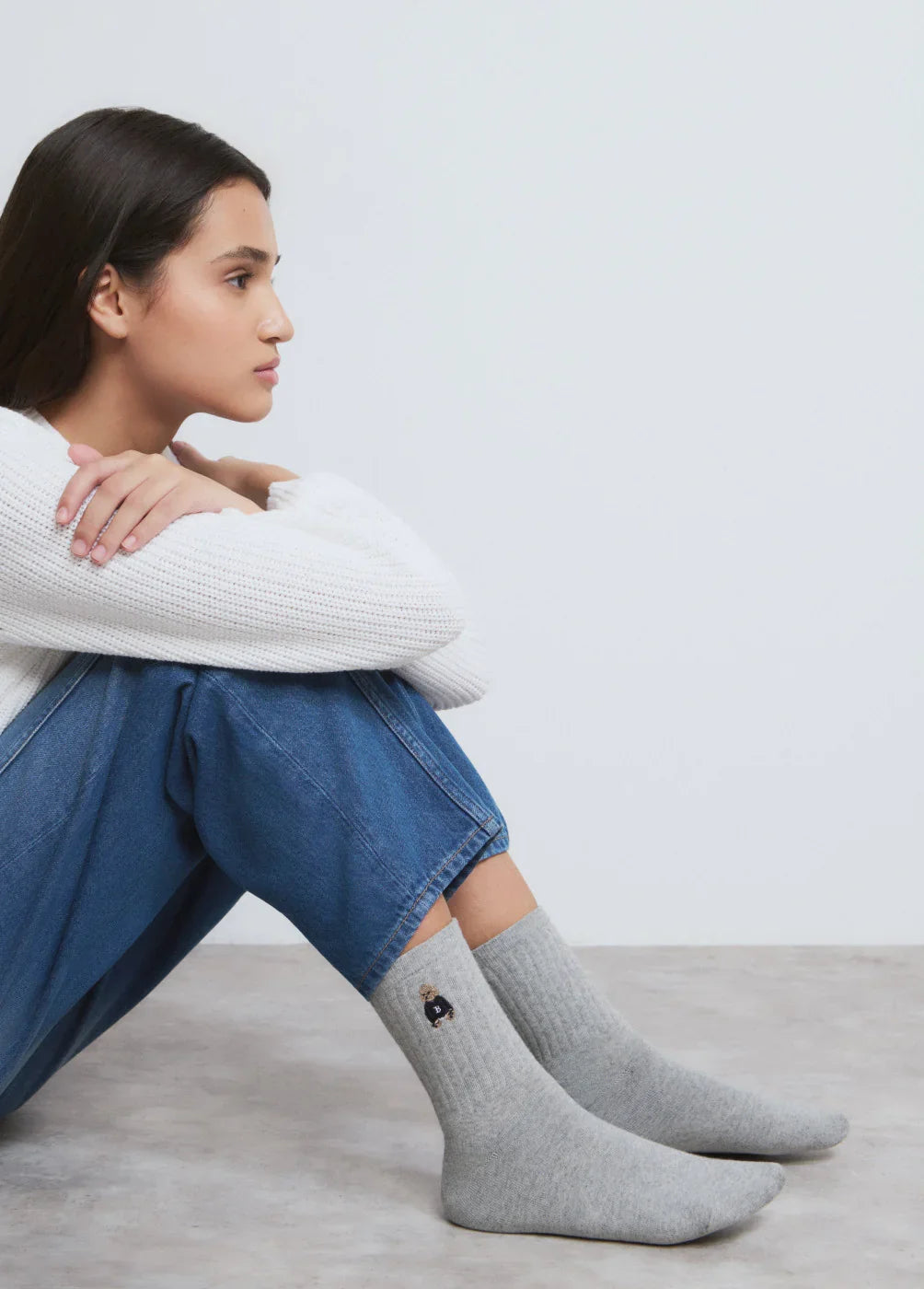אישה יושבת עם סוודר לבן, ג'ינס וגרביים אפורות עם דובי, צילום אופנה לחנות CHOZEN