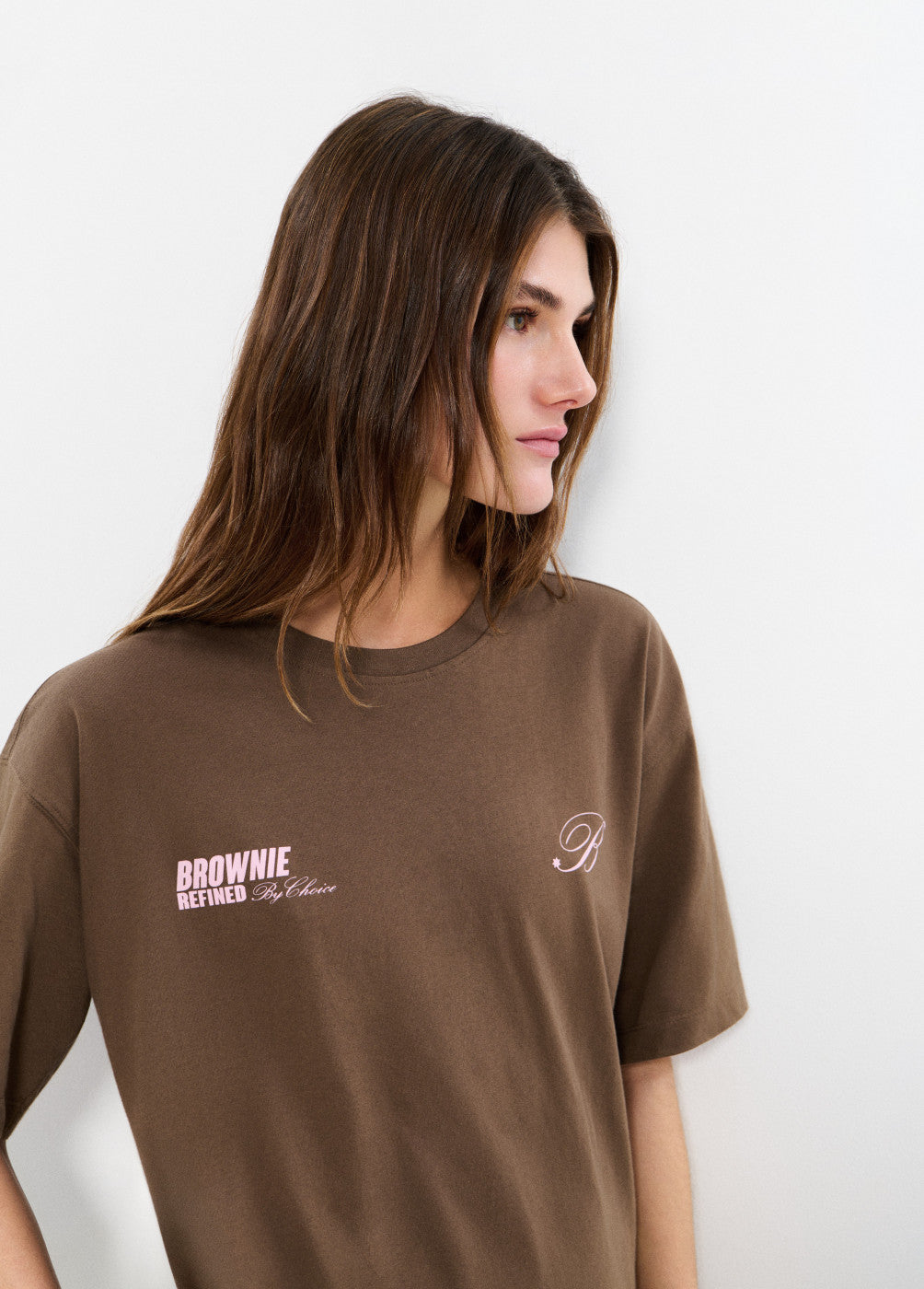 אישה לובשת חולצת טי חומה עם כיתוב BROWNIE REFINED על החזה, רקע לבן