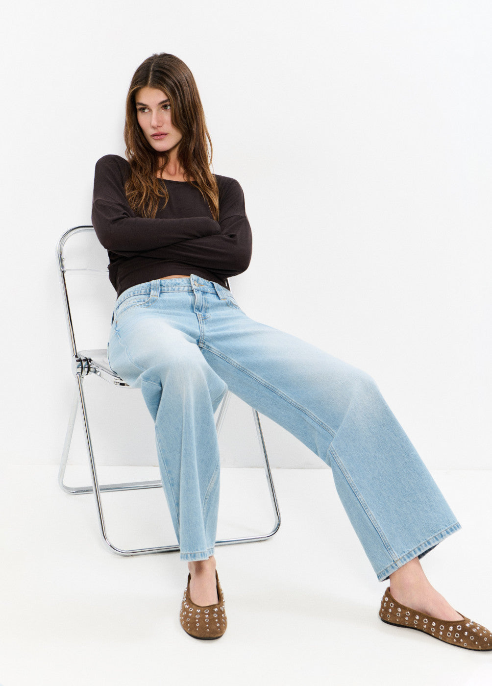 אישה יושבת על כיסא לובשת ג'ינס בגזרה רחבה, חולצה שחורה ונעליים חומות עם ניטים