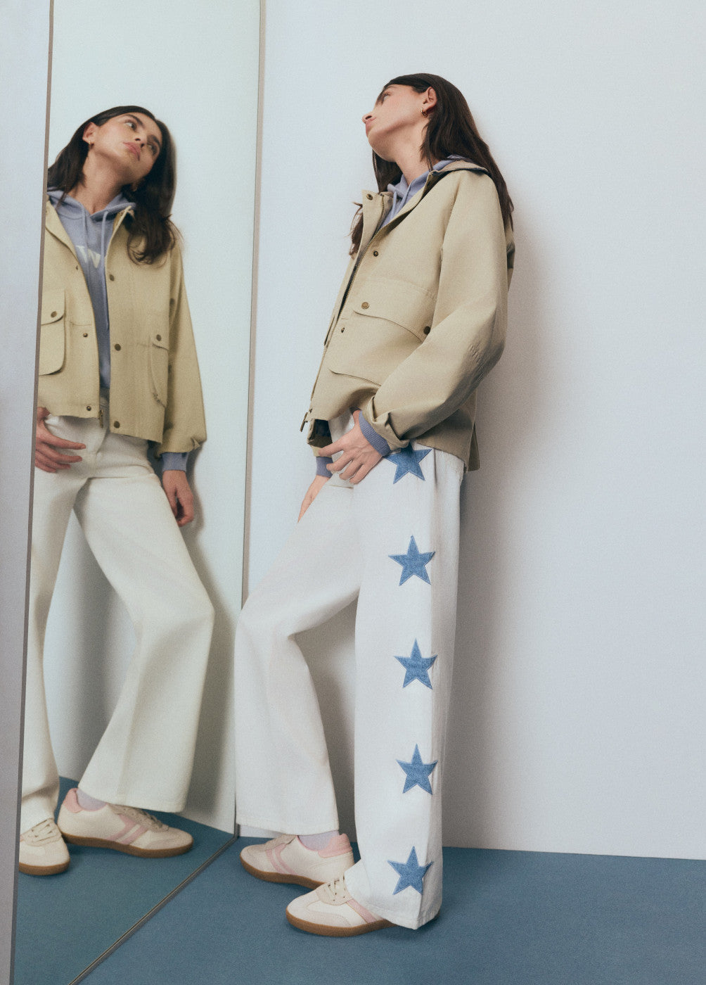 אישה לובשת ג'קט בז' ומכנסיים לבנים עם כוכבים כחולים, עומדת מול מראה