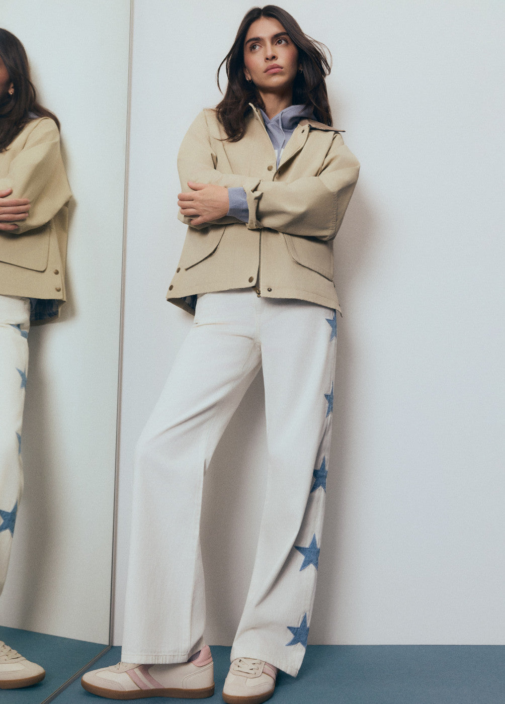 אישה עם ג'קט בז', מכנסיים לבנים עם כוכבים כחולים ונעלי סניקרס אופנתיות עומדת ליד מראה