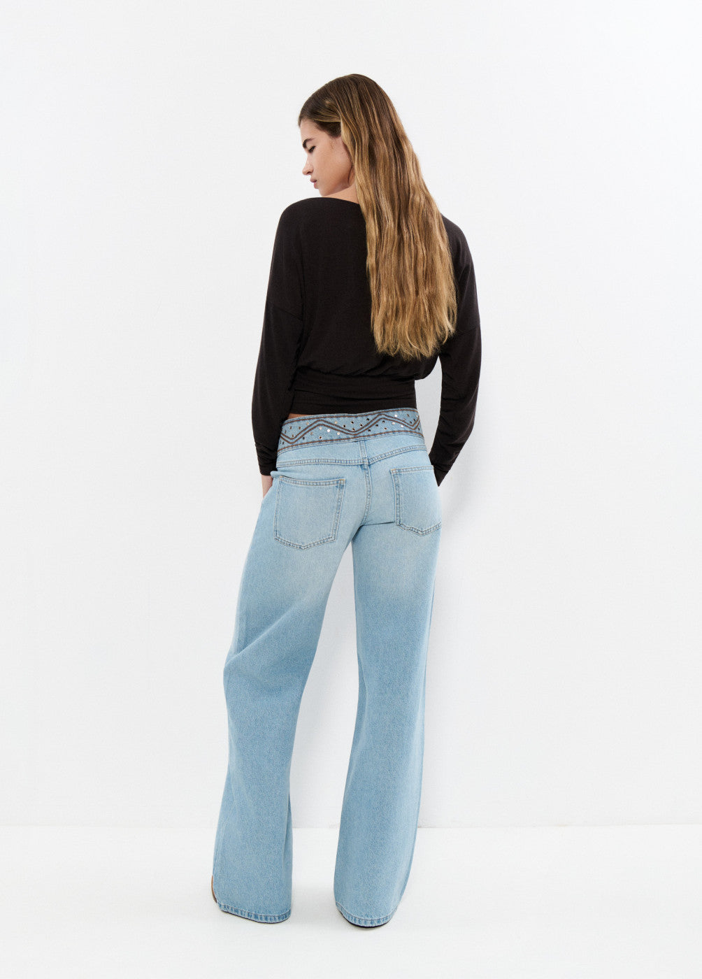 אישה לובשת מכנסי ג'ינס בגזרה גבוהה ורחבה, חולצה שחורה, עומדת עם גב למצלמה