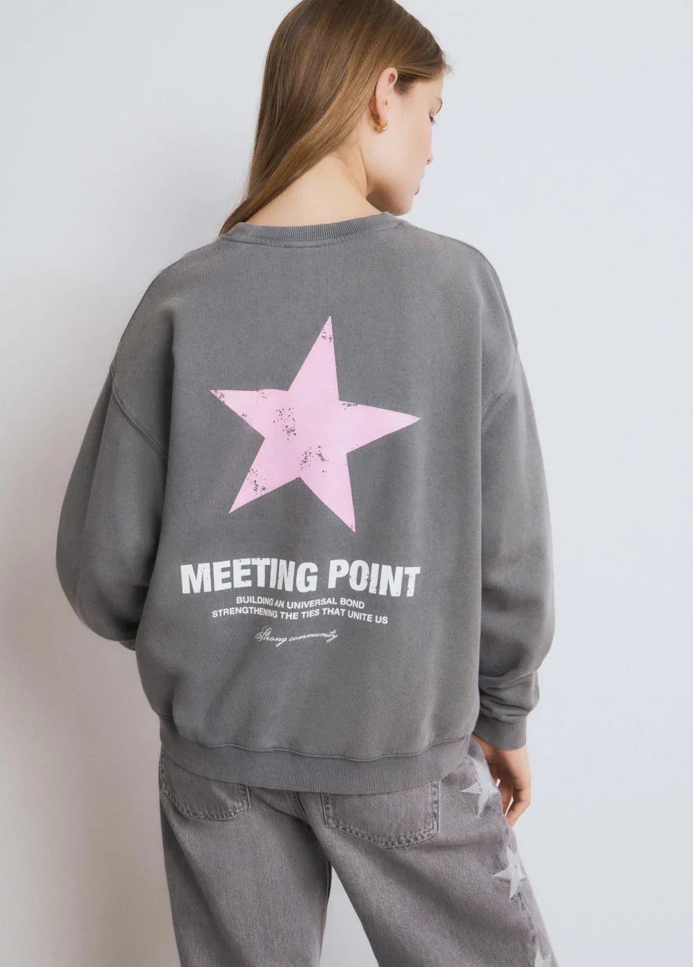 אישה עם סווטשירט אפור של CHOZEN, כוכב ורוד והכיתוב 'MEETING POINT', מכנסי ג׳ינס עם כוכבים