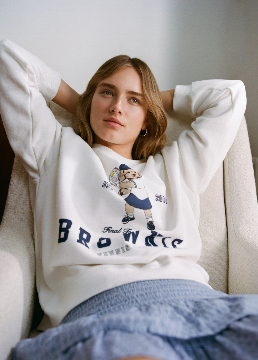 אישה צעירה לובשת סווטשירט לבן עם הדפס של כלב משחק טניס, יושבת על כורסה