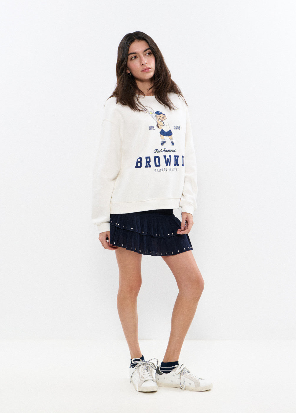 דוגמנית עם סווטשירט לבן בהדפס דמות וטקסט, חצאית פליסה כחולה קצרה ונעלי סניקרס