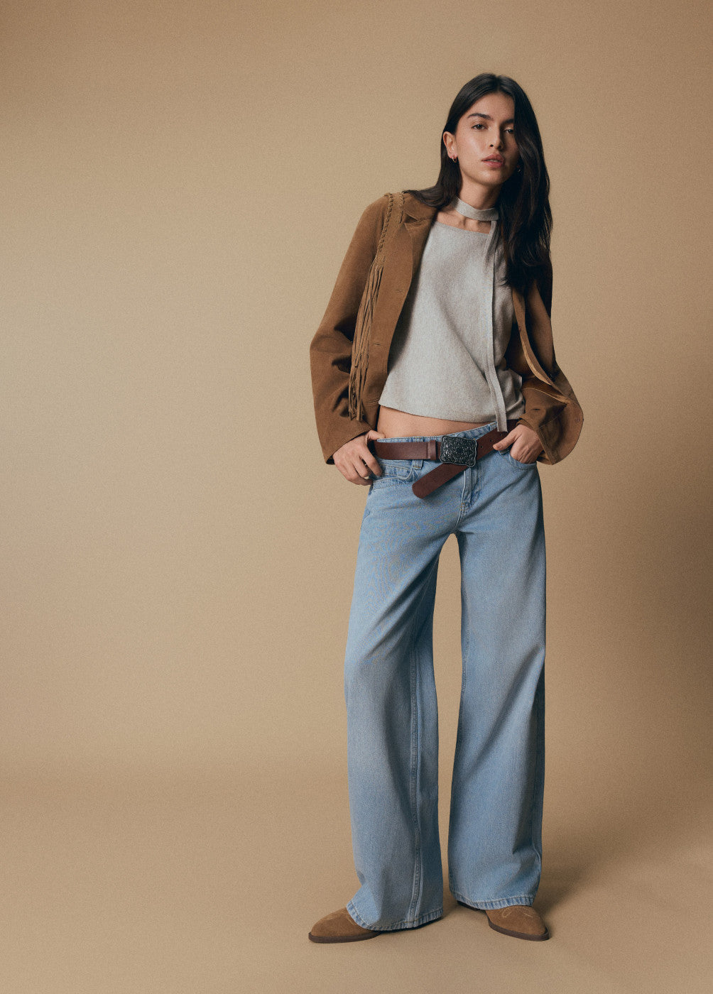 אישה עם ג'ינס רחב, חולצה אפורה, ז'קט חום חגורה חומה ונעליים חומות, רקע בז'