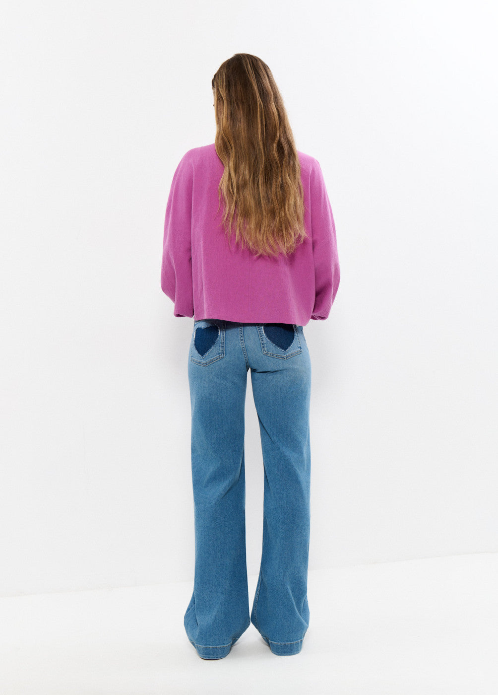 אישה עם שיער ארוך לובשת ג'ינס כחול עם כיסים כהים וחולצה ורודה, מצולמת מאחור