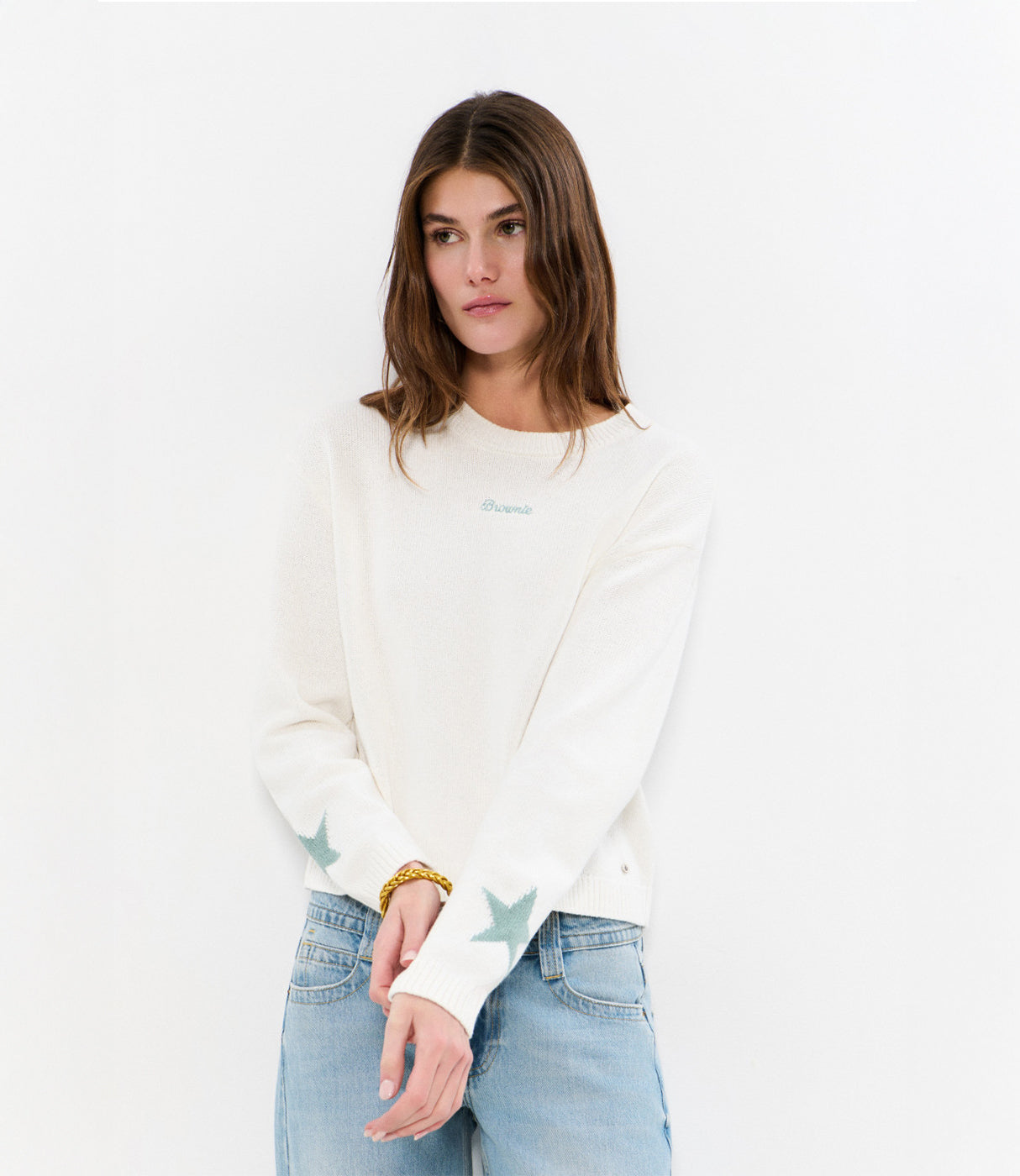 אישה עם סוודר לבן מעוטר כוכבים בירוק על השרוולים, ג'ינס בהיר, רקע לבן