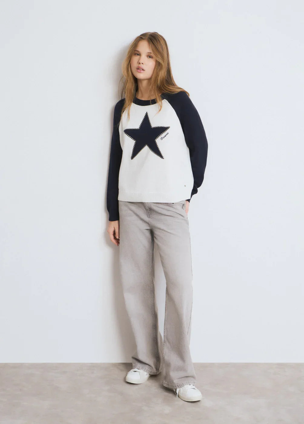 דוגמנית לובשת סוודר לבן עם שרוולים כהים וכוכב שחור מבית CHOZEN, מכנסיים אפורים ונעליים לבנות