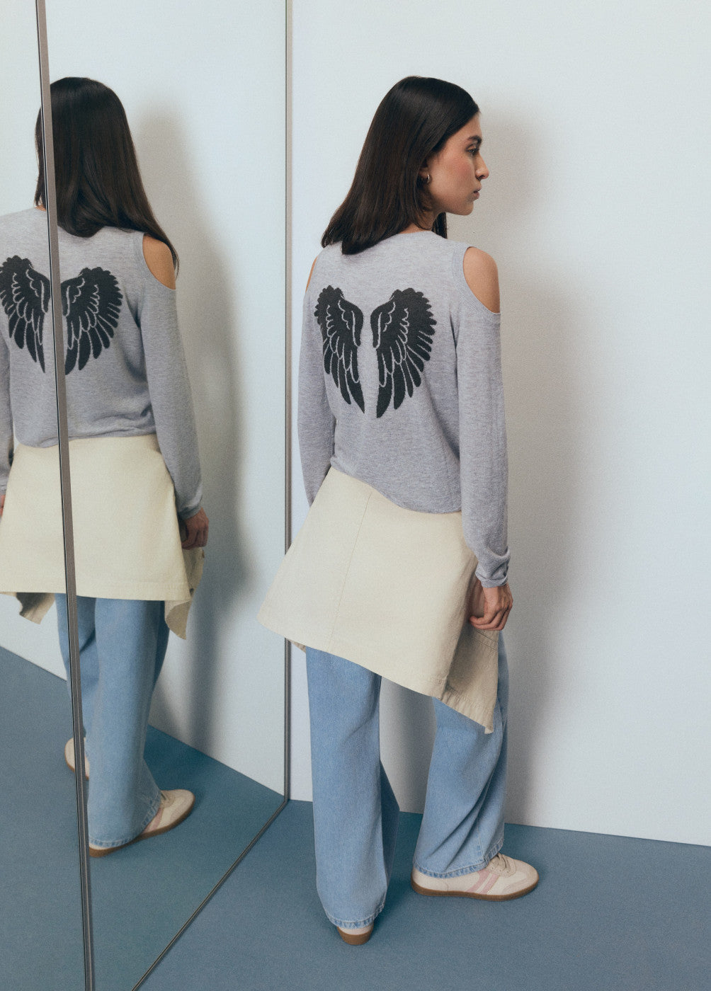 אישה עם סווטשירט אפור מכנפיים שחורות בגב, ג'ינס רחב, נעליים בהירות, עומדת ליד מראה