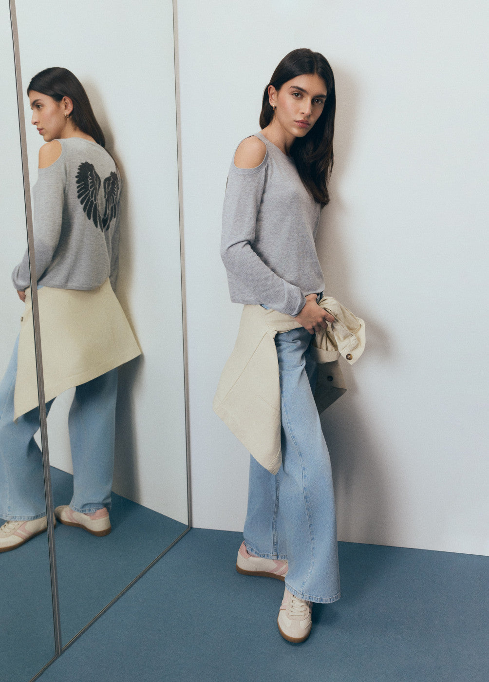 אישה עם סווטשירט אפור בכתפיים חשופות, הדפס כנפיים בגב, ג'ינס רחב ונעליים לבנות