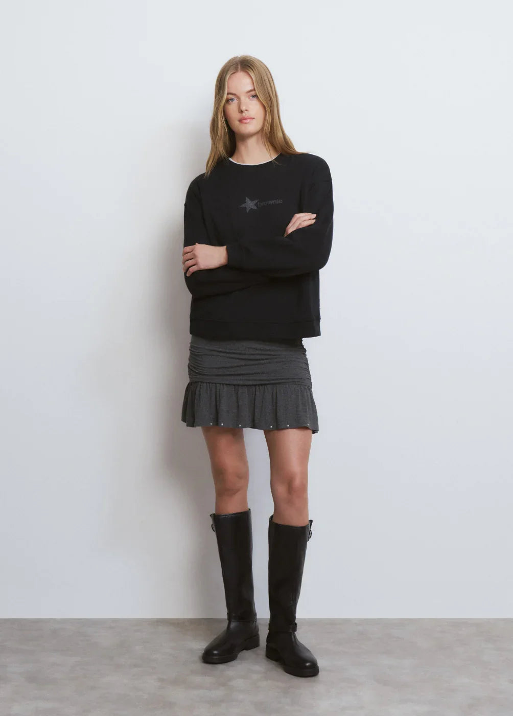 אישה לובשת סווטשירט שחור של CHOZEN, חצאית אפורה קצרה ומגפיים שחורים, סטייל אופנתי