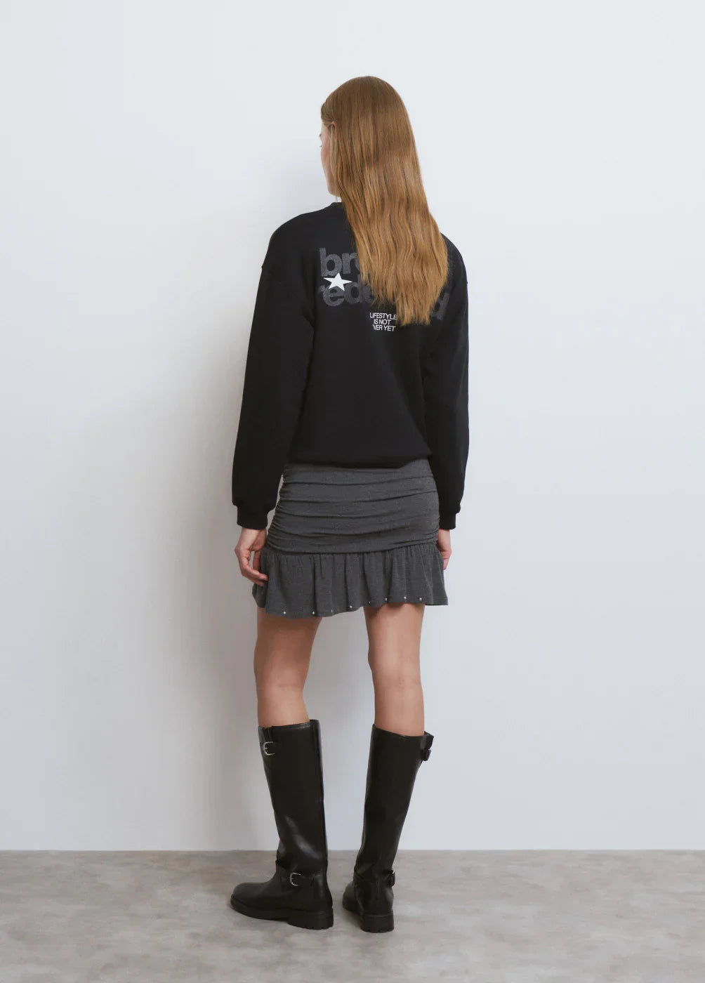 אישה עם חולצת סווטשרט שחורה, חצאית אפורה קצרה ומגפיים שחורים עומדת על רקע לבן, אופנה CHOZEN