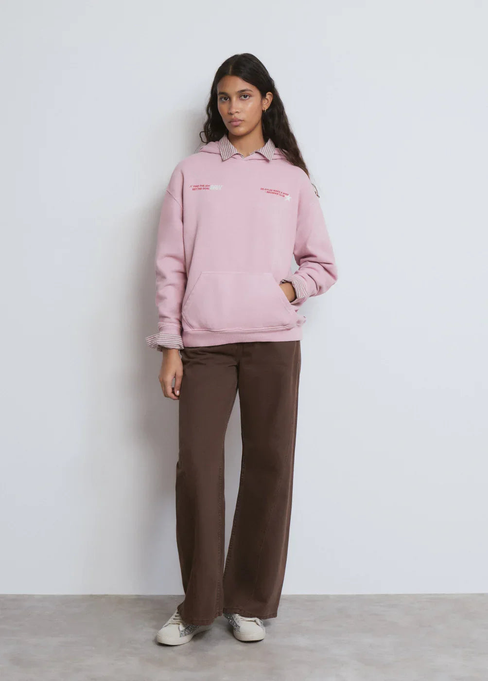 דוגמנית עומדת על רקע לבן לובשת קפוצ'ון ורוד של CHOZEN ומכנסיים חומים, אופנת רחוב