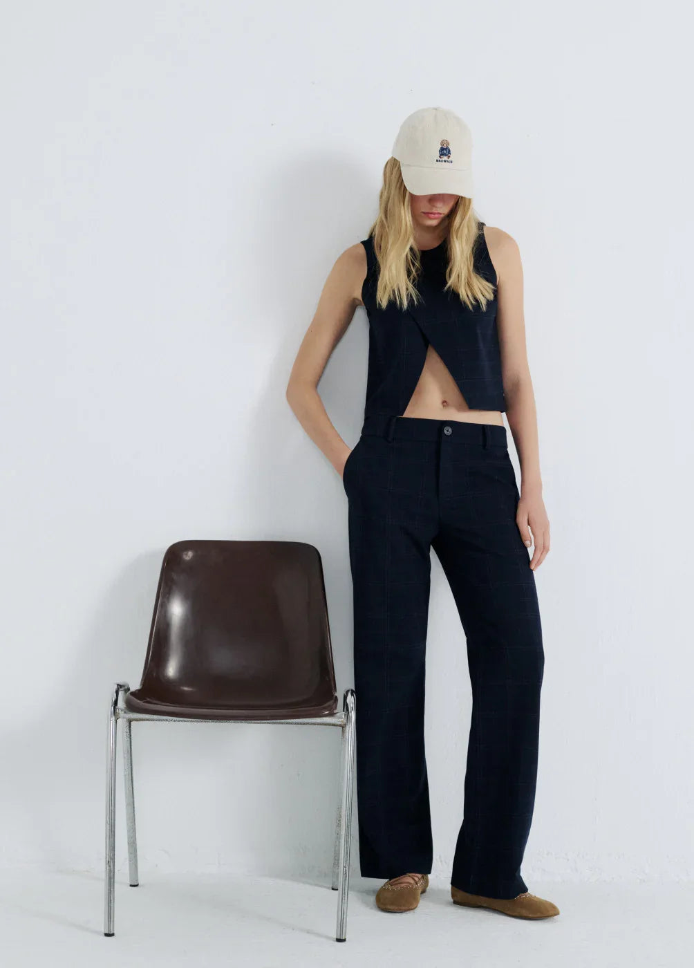 אישה עם גופייה כהה, מכנסיים מחויטים וכובע, עומדת ליד כיסא בחלל מינימליסטי, אופנת CHOZEN