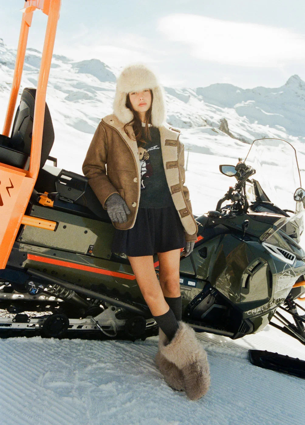 אופנת חורף CHOZEN – דוגמנית במעיל צמר וכובע פרווה ליד אופנוע שלג ברקע נוף שלג הררי