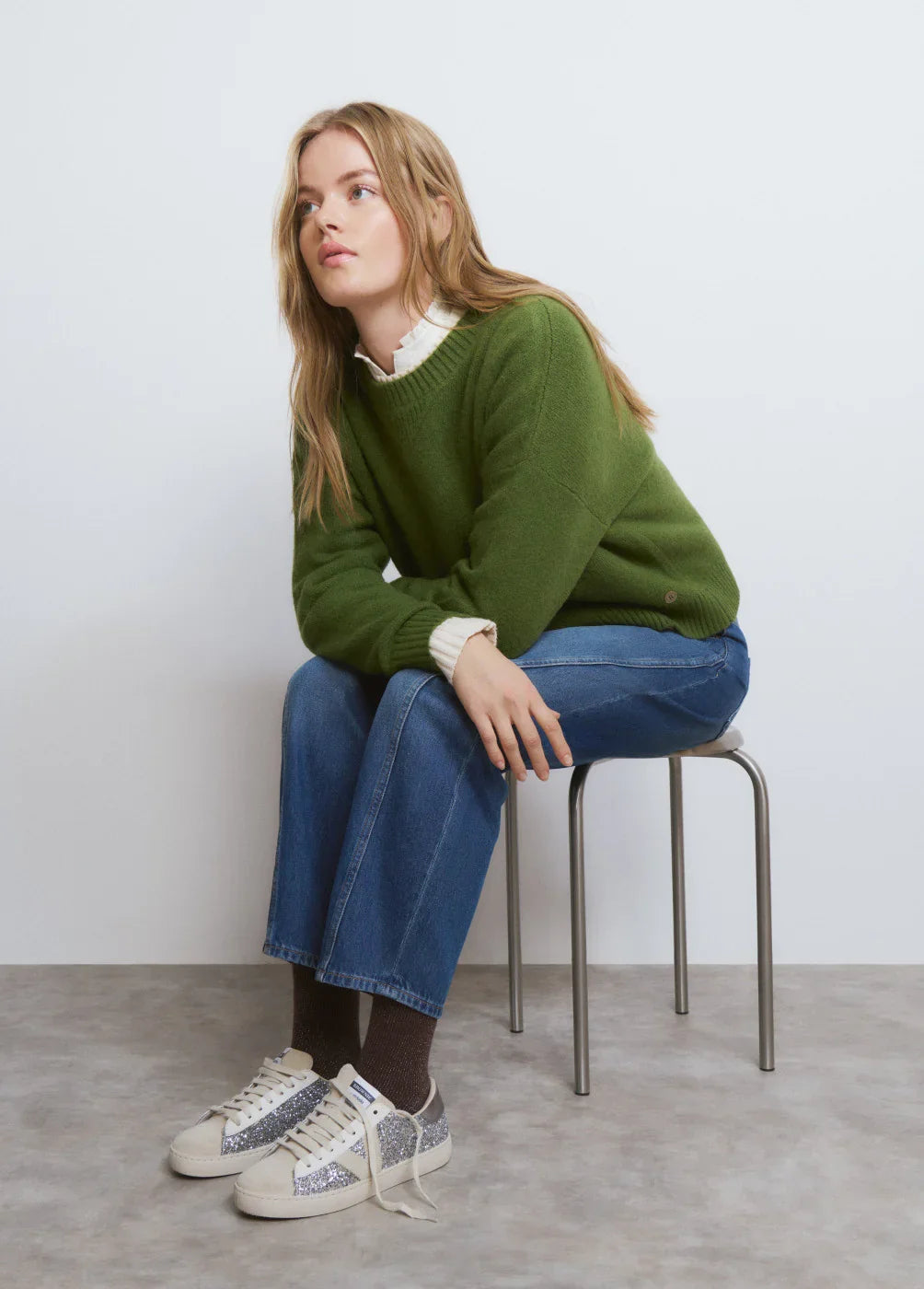 אישה יושבת על כיסא, לובשת סוודר ירוק, ג'ינס ונעלי סניקרס נוצצות, אופנת CHOZEN