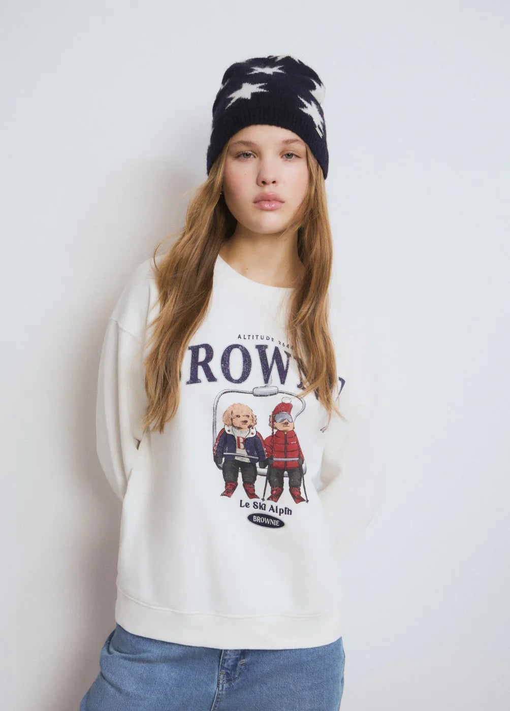 דוגמנית עם סווטשירט לבן עם הדפסת דובי ומכנסי ג'ינס, חבושה בכובע חורף שחור עם כוכבים, אופנה CHOZEN