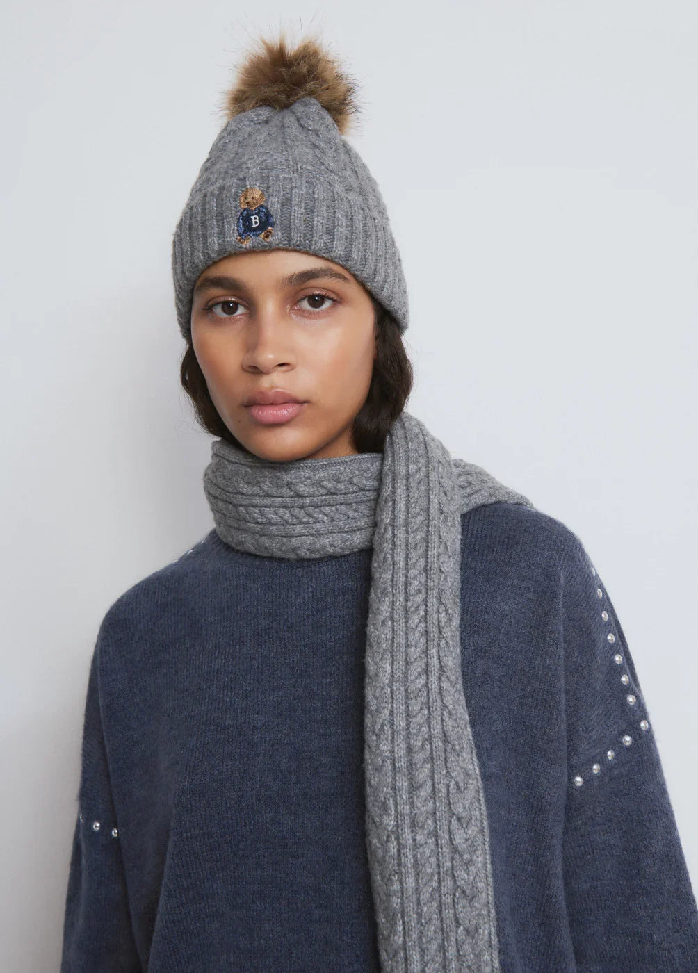 אישה עם כובע סרוג אפור עם פונפון ודובי, צעיף סרוג באפור, סוודר כחול, אופנת חורף CHOZEN