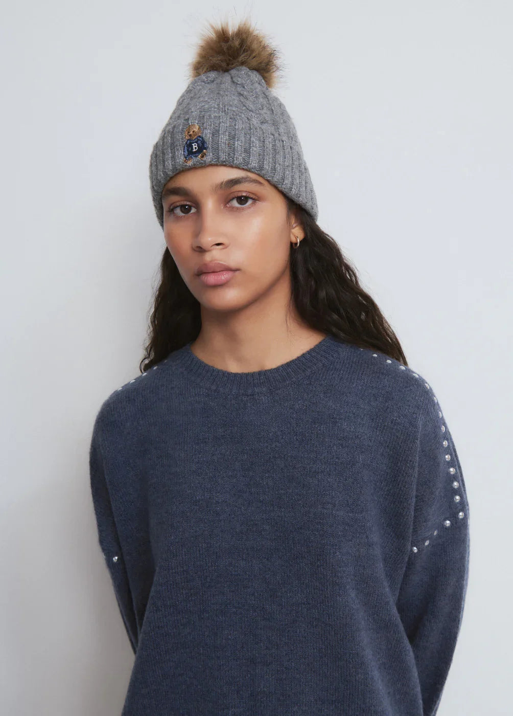 אישה לובשת כובע צמר אפור עם פונפון חום ודובי רקום, וסוודר כחול כהה, מבית CHOZEN