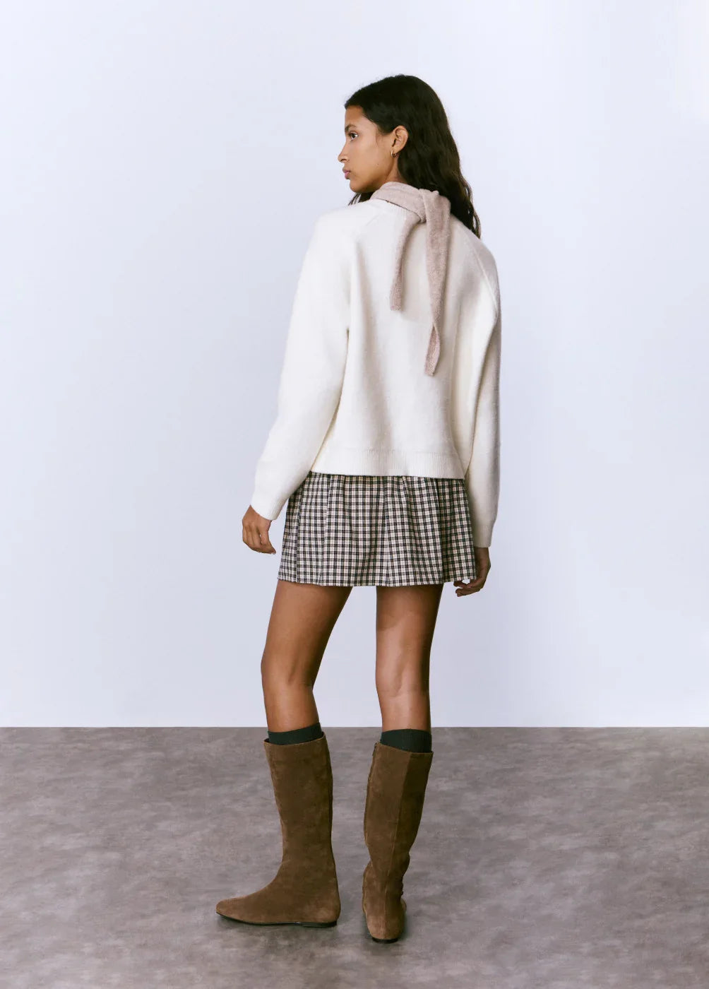 דוגמנית עם סוודר לבן, חצאית משובצת ומגפיים חומים מציגה אופנת חורף CHOZEN